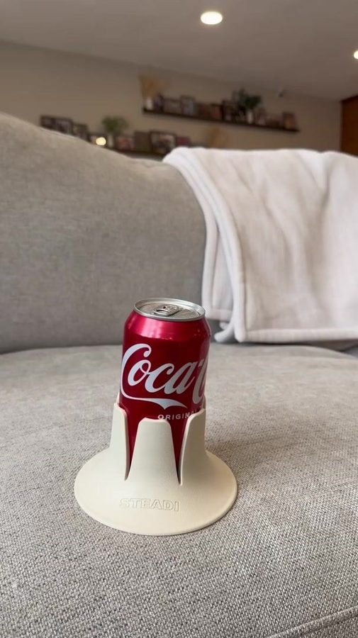 A full can standing in a bone-coloured Steadi on a grey couch cushion, a living room behind it.