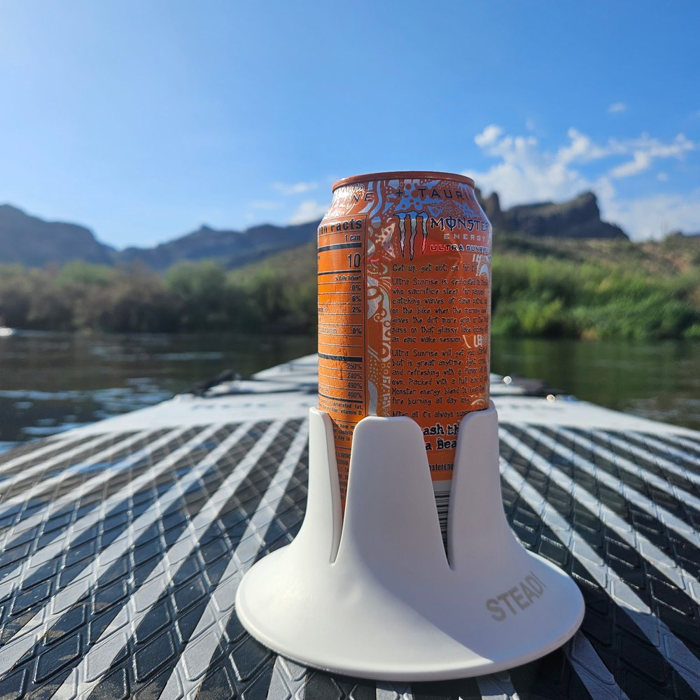 A white Steadi holding an orange energy drink can on the textured deck pad of a paddleboard, a desert river and red rock ridgeline behind it.