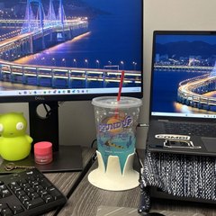 A white/marbled drink base holding a clear cup of blue drink with a red straw on a grey wood-look home-office desk, between a large monitor, a laptop on a stand, a split keyboard and a mouse pad