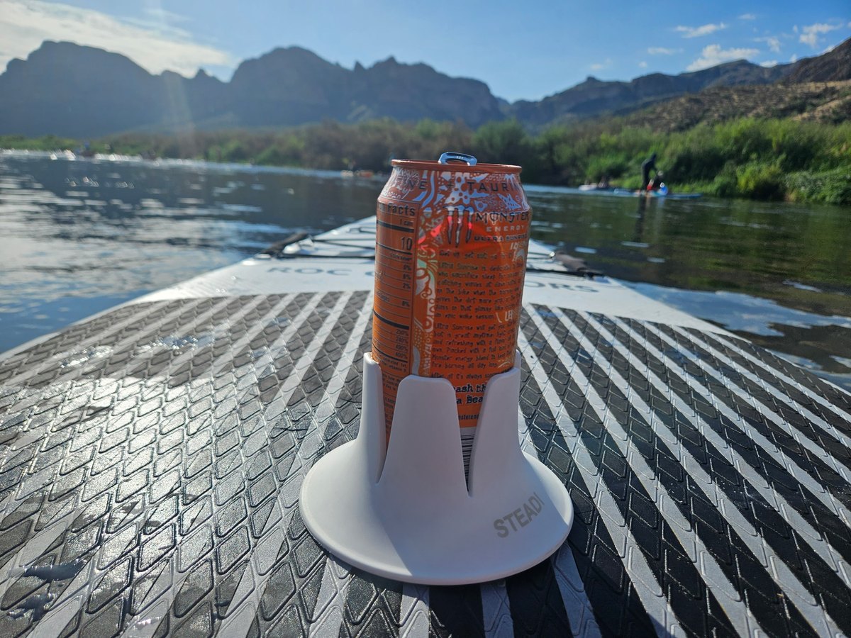 A white Steadi holding an orange can on a paddleboard deck, another paddler downriver and a red rock ridgeline across the horizon.