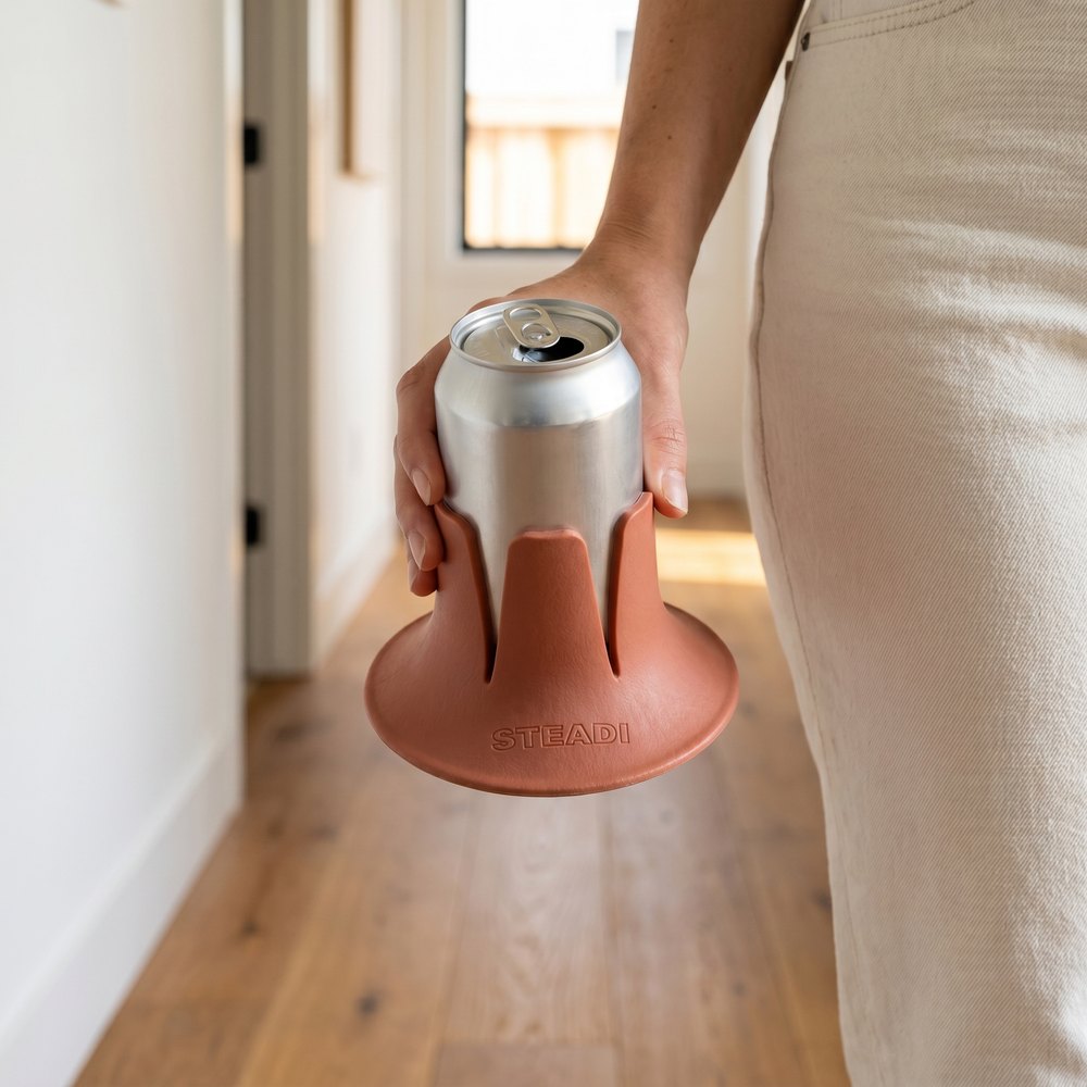 A hand carrying a terracotta Steadi with a full can seated in it, held by the base at waist height.